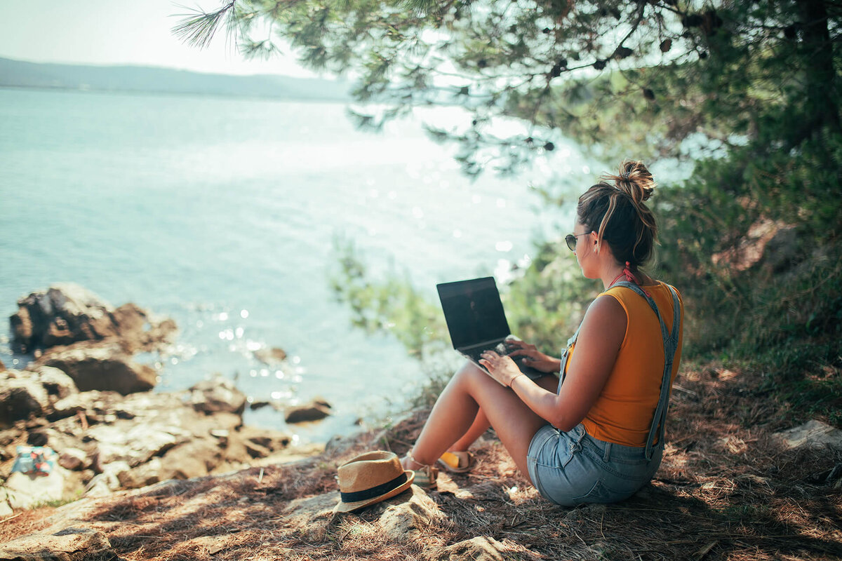 Frau sitzt mit Laptop unter Baum am See - Volunteering mit Volunta Erfahrungsberichte